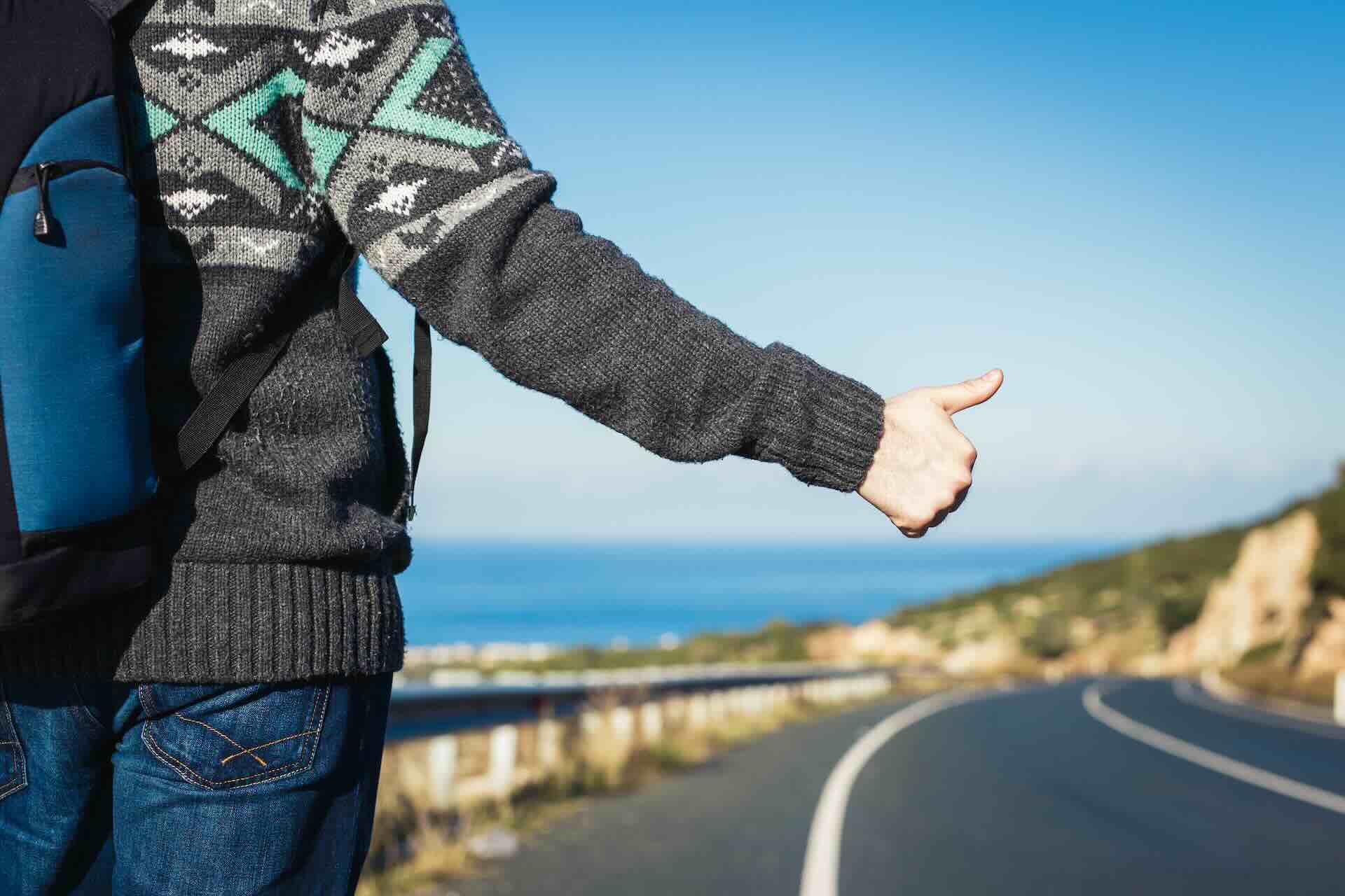 A hitchhiker next to the highway with her thumb sticking up near the coastline.